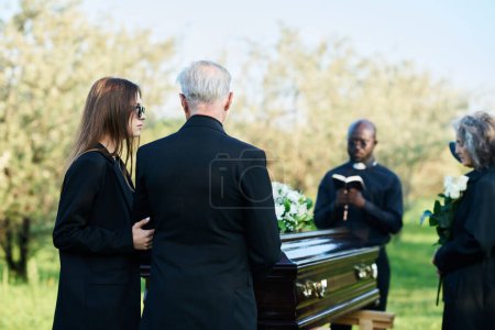 Mature man and his daughter in mourning attire standing in front of coffin with closed lid and group of people during funeral service with priest