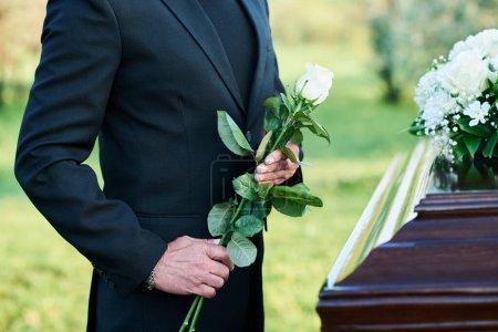 Schnappschuss eines reifen Witwers im schwarzen Anzug, der frische weiße Rosen hält, während er am Sarg mit dem Leichnam seiner Frau bei der Beerdigung steht