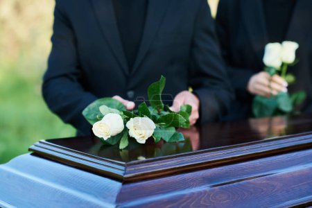 Konzentrieren Sie sich auf zwei frische weiße Rosen, die von einem trauernden reifen Mann im schwarzen Anzug auf den Sargdeckel gelegt werden, der gegen eine junge Frau mit Blumen steht