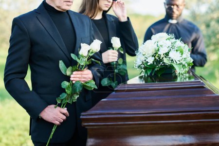 Cropped shot of mature man with two white roses standing by coffin with bunch of fresh chrysanthemums on top with his daughter on background