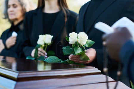 Focus on two fresh white roses held by mourning man in black suit during funeral service while standing by coffin against his daughter and wife