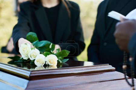 Mujer joven en traje negro poniendo cuatro rosas blancas frescas en la tapa cerrada del ataúd mientras está de pie junto a su padre y sacerdote leyendo la Biblia