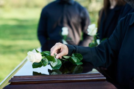 Main de femme mûre en tenue de deuil mettant rose blanche sur le dessus du couvercle fermé du cercueil tout en se tenant devant la caméra contre d'autres personnes