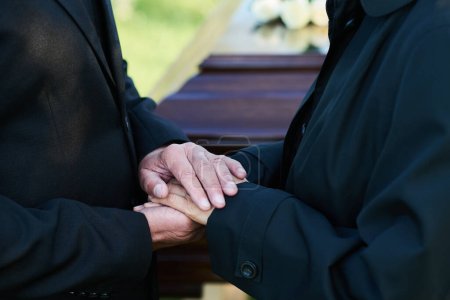 Close-up of hands of mature grieving man on those of his mourning wife in black attire standing in front of camera against closed coffin