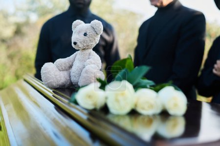 Photo for Focus on grey teddybear sitting on top of closed coffin lid with bunch of fresh white roses standing in front of camera against grieving people - Royalty Free Image