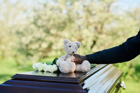 Hand of young African American man in black shirt putting teddybear on top of coffin with his dead bride or wife while saying last goodbye