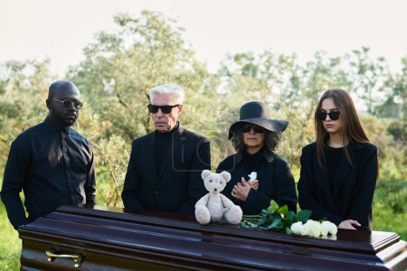 Téléchargez les photos : Groupe de personnes interculturelles en deuil dans des vêtements de deuil réunis par cercueil avec ours en peluche et roses blanches fraîches sur le dessus du couvercle fermé - en image libre de droit