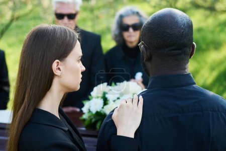 Téléchargez les photos : Vue arrière de la jeune femme en deuil tenant la main sur l'épaule de son mari en costume de deuil tandis que les deux debout devant le cercueil - en image libre de droit