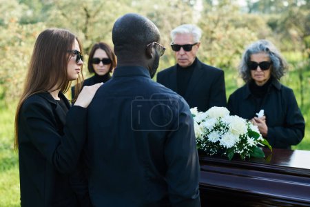 Foto de Vista posterior de la joven mujer en traje de luto apoyando a su marido en el funeral mientras ambos de pie frente al ataúd con ramo de flores blancas - Imagen libre de derechos