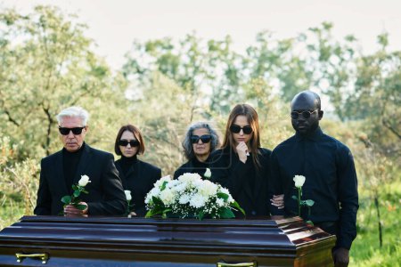 Téléchargez les photos : Groupe de personnes en deuil interculturel portant des vêtements de deuil et des lunettes de soleil tenant des roses blanches debout devant un cercueil avec couvercle fermé - en image libre de droit