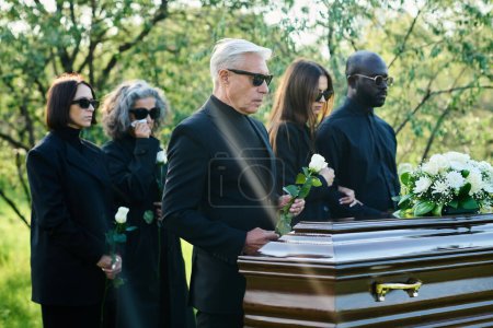 Téléchargez les photos : Des amis et des parents en deuil d'une personne décédée avec des roses blanches debout près d'un cercueil avec des chrysanthèmes sur le dessus du couvercle fermé - en image libre de droit