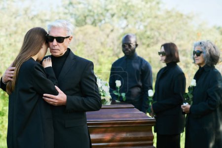 Trauernder reifer Mann im schwarzen Anzug umarmt seine trauernde Tochter und drückt gleichzeitig sein Mitgefühl für den Verlust eines lieben Freundes oder Familienmitglieds aus