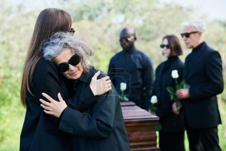 Téléchargez les photos : Jeune femme en deuil faisant un câlin à sa mère en deuil avec des lunettes de soleil pleurant sur son épaule tout en se tenant contre le cercueil et le groupe de personnes - en image libre de droit