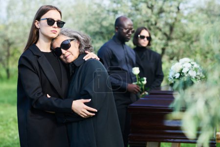 Mature grieving woman in sunglasses and mourning attire keeping head on shoulder of her daughter while both standing in front of camera