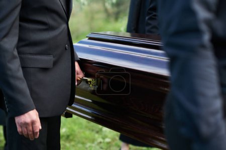 Focus on man in black suit holding by handle of wooden coffin while carrying it together with other people after farewell ceremony