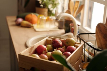 Téléchargez les photos : Mettre l'accent sur la boîte en bois avec des pommes mûres rouges debout devant la caméra à côté du panier avec des serviettes propres roulées contre l'évier sur le comptoir de la cuisine - en image libre de droit