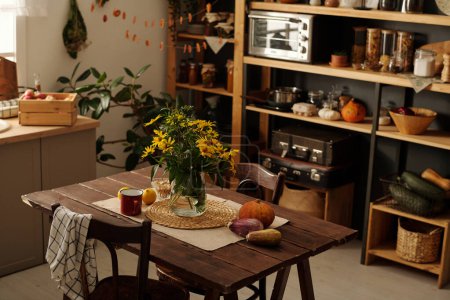 Table en bois avec bouquet de fleurs sauvages dans un vase, légumes, tasse et deux chaises debout dans le centre de la cuisine spacieuse de la maison de campagne
