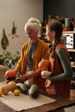 Photo for Senior woman with white hair and her granddaughter standing by wooden table in the kitchen and choosing fresh vegetables for cooking stew - Royalty Free Image