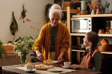 Téléchargez les photos : Femme âgée mettant morceau de tarte aux pommes maison sur l'assiette de sa mignonne petite-fille assise à table dans la cuisine et ayant une tisane - en image libre de droit