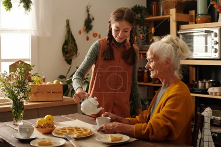 Photo for Happy girl in casualwear pouring herbal tea into cup of her grandmother sitting by wooden table served with appetizing homemade apple pie - Royalty Free Image