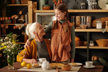 Téléchargez les photos : Grand-mère joyeuse embrassant sa petite-fille mignonne et la regardant tout en étant assis à table servi avec de la nourriture maison et prendre le thé - en image libre de droit