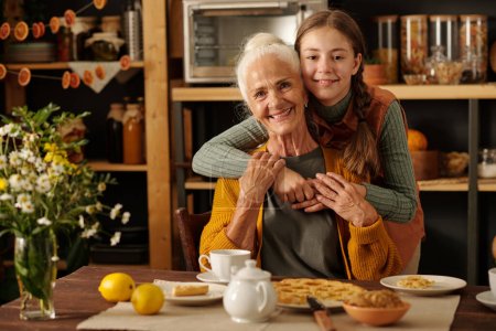 Nettes fröhliches Mädchen mit Zöpfen umarmt ihre Großmutter am Tisch sitzend mit hausgemachtem Apfelkuchen und Tee, während beide in die Kamera schauen