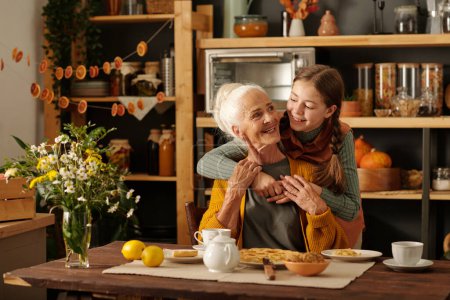 Téléchargez les photos : Jolie fille embrassant sa grand-mère aînée joyeuse assise à côté d'une table en bois avec tarte aux pommes maison appétissante, citrons et tisane - en image libre de droit