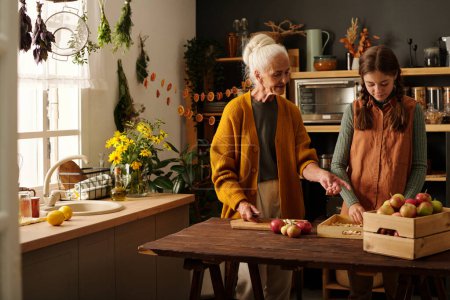 Foto de Mujer anciana con el pelo blanco de pie junto a la mesa de su nieta ayudando con la preparación de frutas y verduras frescas para el invierno - Imagen libre de derechos