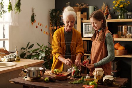 Téléchargez les photos : Jolie fille avec des tomates fraîches aidant sa grand-mère à préparer des cornichons pour l'hiver tout en regardant la femme âgée mettre des légumes dans un pot - en image libre de droit