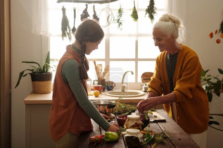 Photo for Side view of happy grandmother and granddaughter making pickles together while senior woman putting fresh vegetables into jar - Royalty Free Image