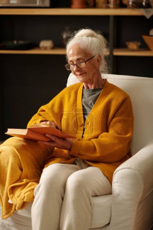 Téléchargez les photos : Femme âgée sérieuse en cardigan jaune et pantalon blanc livre de lecture tout en étant assis dans un grand fauteuil confortable à loisir dans une maison de campagne - en image libre de droit