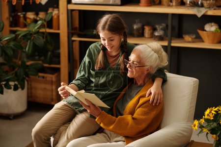 Cute girl embracing her grandmother while sitting next to her in armchair and both reading book while enjoying rest in country house in autumn