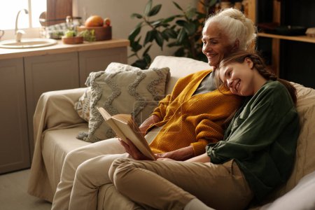 Téléchargez les photos : Joyeux fille mignonne gardant la tête sur l'épaule de sa grand-mère souriante avec livre ouvert tout en étant assis sur le canapé à côté d'elle et profiter des loisirs - en image libre de droit
