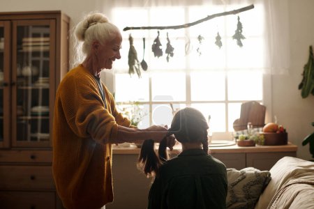 Téléchargez les photos : Vue latérale de heureuse femme âgée aux cheveux blancs tresses à tresses à sa petite-fille tout en se tenant devant elle contre la fenêtre de la cuisine - en image libre de droit