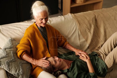 Foto de Chica feliz con coletas disfrutando de pasar tiempo con su abuela mientras se relaja en el sofá y mantener la cabeza en las rodillas de la abuela - Imagen libre de derechos