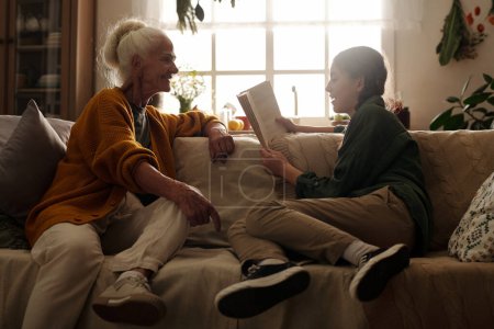Foto de Feliz anciana en ropa casual sentada en el sofá frente a su nieta leyendo cuento o novela en voz alta el fin de semana de otoño - Imagen libre de derechos