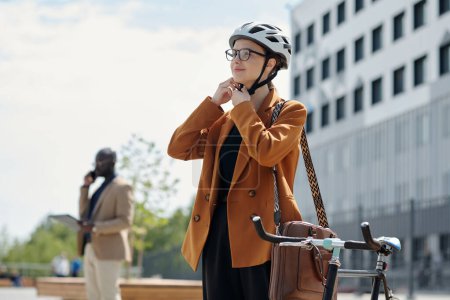 Téléchargez les photos : Heureuse jeune employée attachant sa ceinture de casque de protection tout en se tenant devant sa caméra près de son vélo en milieu urbain - en image libre de droit