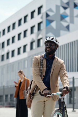 Foto de Feliz joven empresario afroamericano en casco de ciclismo de pie junto a su bicicleta contra la arquitectura moderna mientras va a trabajar - Imagen libre de derechos