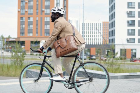 Téléchargez les photos : Vue latérale de l'entrepreneur afro-américain masculin en casque de protection et vêtements de luxe silencieux chevauchant pour travailler à vélo en milieu urbain - en image libre de droit