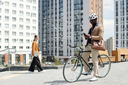 Foto de Joven empresario masculino sonriente en casco de seguridad sentado en bicicleta y desplazándose a través de la información en línea en el teléfono móvil en la calle - Imagen libre de derechos