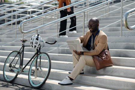 Foto de Joven empresario exitoso en ropa formal mirando a la pantalla del ordenador portátil mientras está sentado en la escalera en el entorno urbano y la creación de redes - Imagen libre de derechos