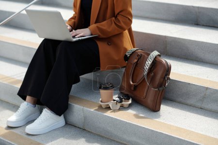 Cropped shot of young stylish female digital nomad typing on laptop keyboard while sitting on grey concrete staircase in front of camera