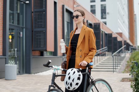 Photo for Young modern female entrepreneur with bottle of water and bicycle with protective helmet standing in front of camera in urban environment - Royalty Free Image