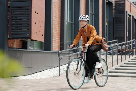 Téléchargez les photos : Jeune femme en tenue de cérémonie et casque de sécurité assis sur la bicyclette et à cheval à la maison du travail tout en se déplaçant le long du bâtiment moderne - en image libre de droit