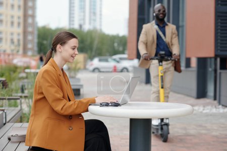 Photo for Focus on young successful businesswoman or student sitting in front of laptop by table in urban environment and typing on keyboard - Royalty Free Image