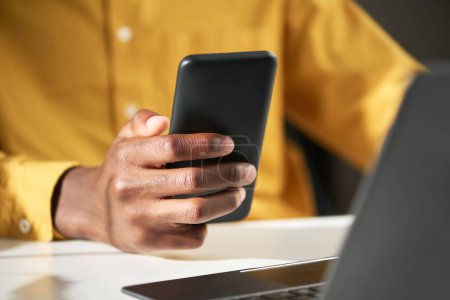 Téléchargez les photos : Focus sur le téléphone portable en main du jeune gestionnaire afro-américain ou du directeur général envoyant des textos devant un ordinateur portable assis à côté du bureau - en image libre de droit