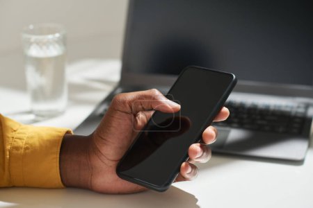 Téléchargez les photos : Main de jeune travailleur de bureau afro-américain touchant l'écran du smartphone avec le pouce tout en étant assis par bureau et en cherchant sur Internet - en image libre de droit
