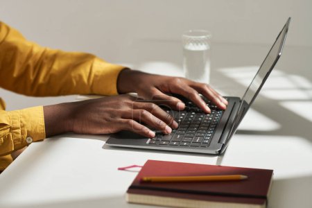 Téléchargez les photos : Les mains du jeune analyste afro-américain masculin ou courtier appuyant sur les touches du clavier de l'ordinateur portable tout en étant assis par bureau dans le bureau et de travailler sur le projet - en image libre de droit