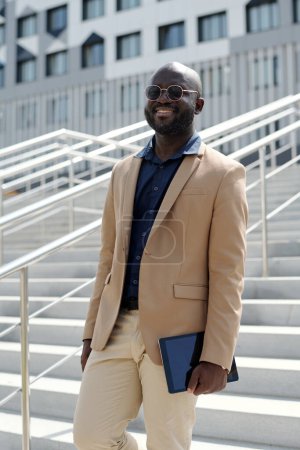 Téléchargez les photos : Jeune solpreneur afro-américain à succès en tenue de luxe silencieuse et lunettes regardant la caméra tout en se tenant debout contre l'escalier - en image libre de droit