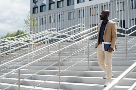 Foto de Joven solopreneur afroamericano serio con documentos financieros y tableta en la mano caminando escaleras abajo contra la construcción - Imagen libre de derechos